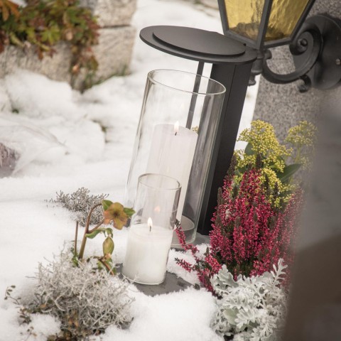Zwei weiße Kerzen in Glasbehältern neben einer schwarzen Wandlaterne und winterlichen Pflanzen im Schnee.